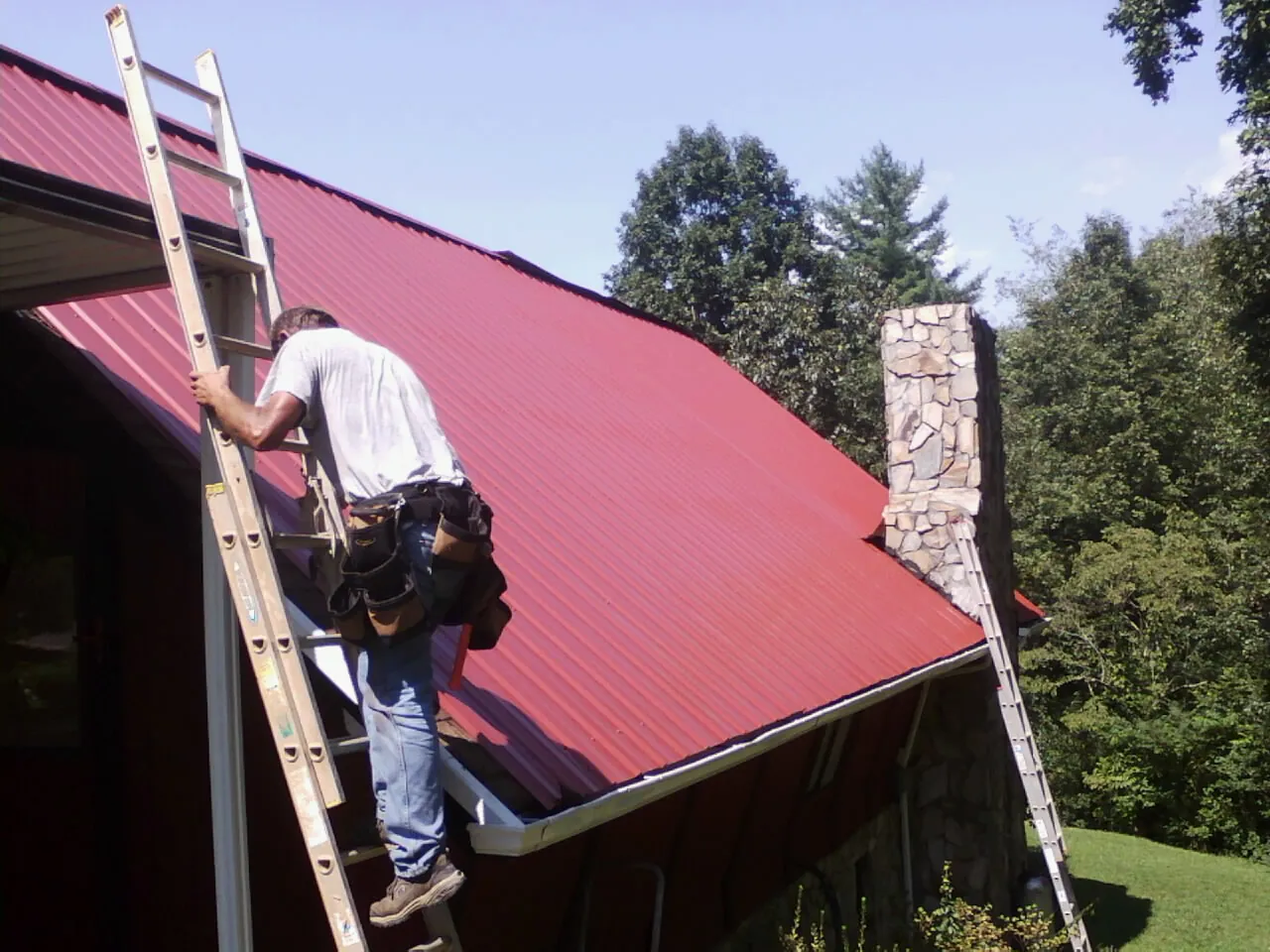 Roofer on a ladder beside a red metal roof and stone chimney — Soffit & Fascia Repair by Roofing Brothers in Shafer